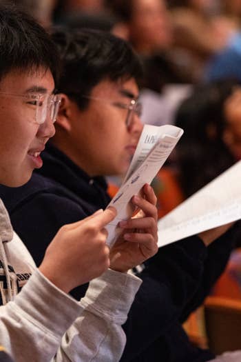 Students wearing glasses read printed worksheets in a classroom with colorful, blurred background lights.