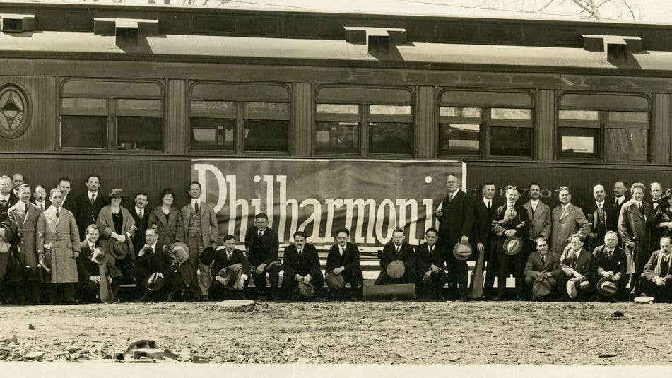 Group posed in front of a train car with “Philharmonic” banner and men holding hats and briefcases