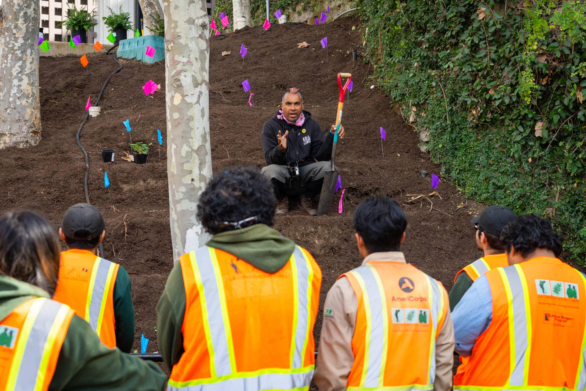 Man seated in dirt garden bed, holding shovel and speaking to workers in orange vests; colorful flag tags fly overhead.
