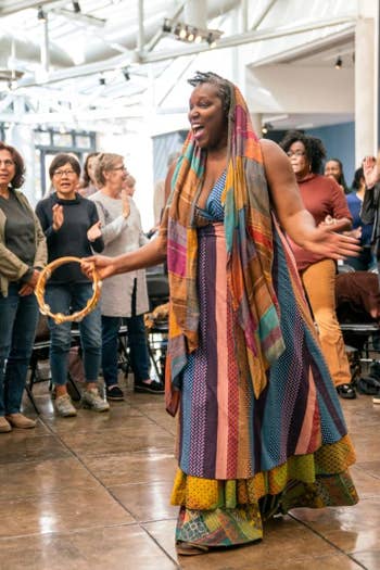 Woman in colorful dress leads a group clap and celebration in a brightly lit indoor event hall