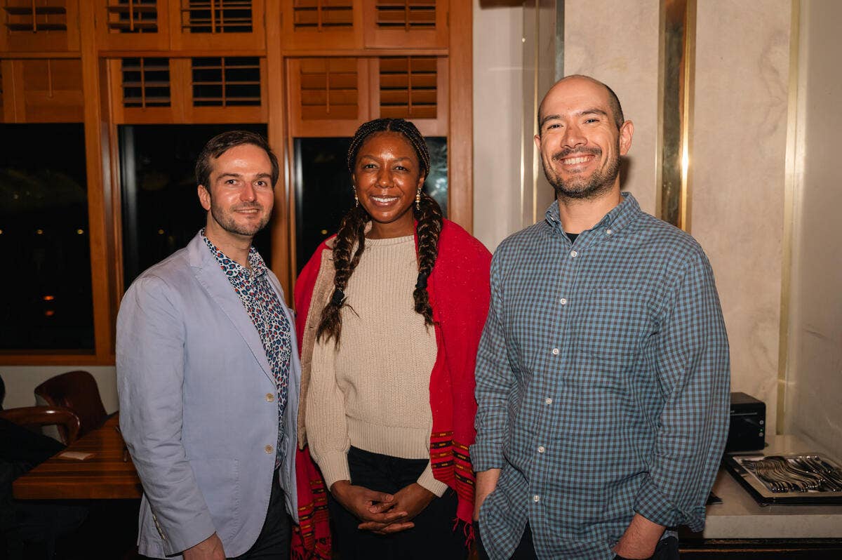 Three smiling people pose indoors, with wooden building windows behind them.
