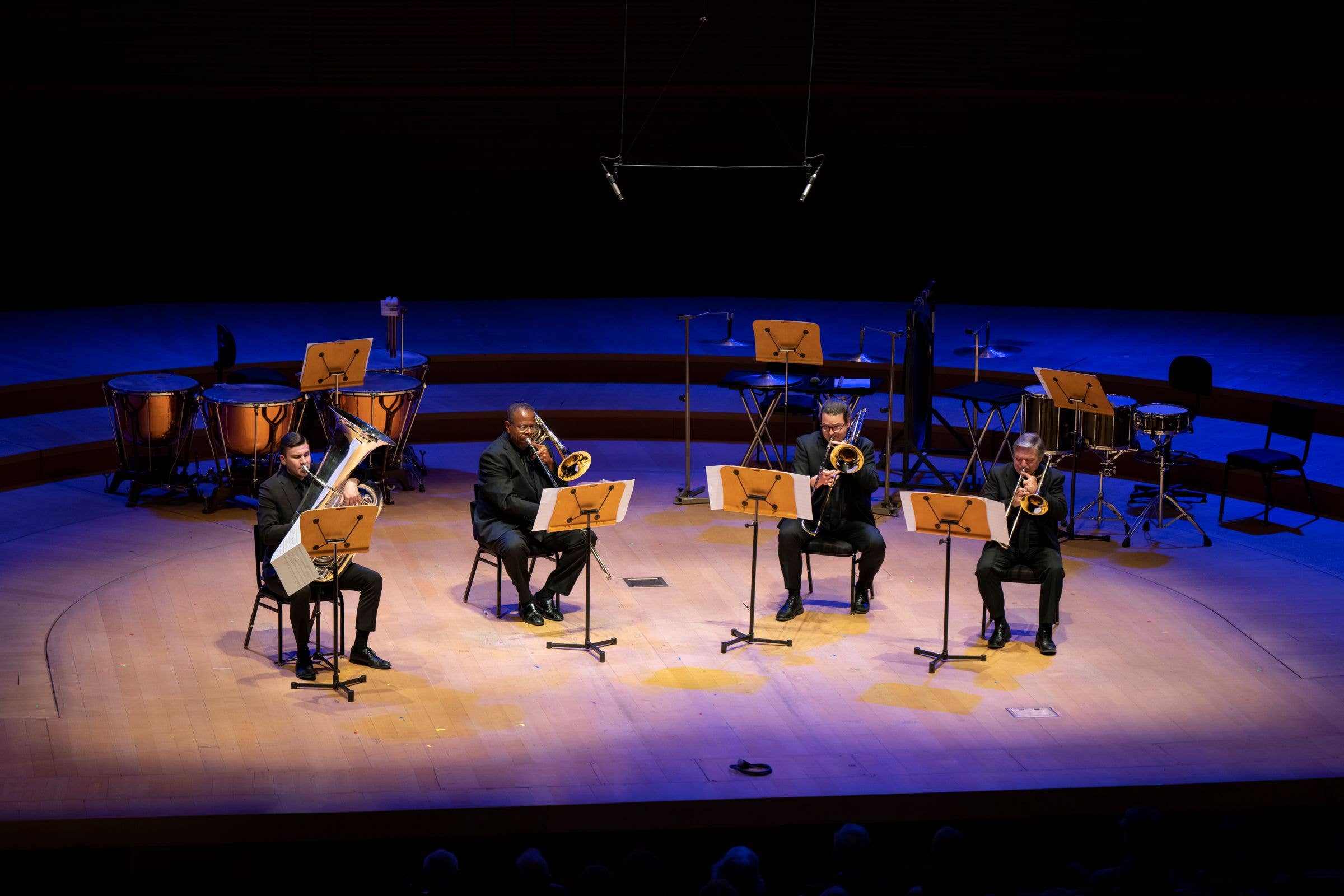 Five seated musicians perform with brass instruments on a concert stage, backed by drums and music stands under blue lighting