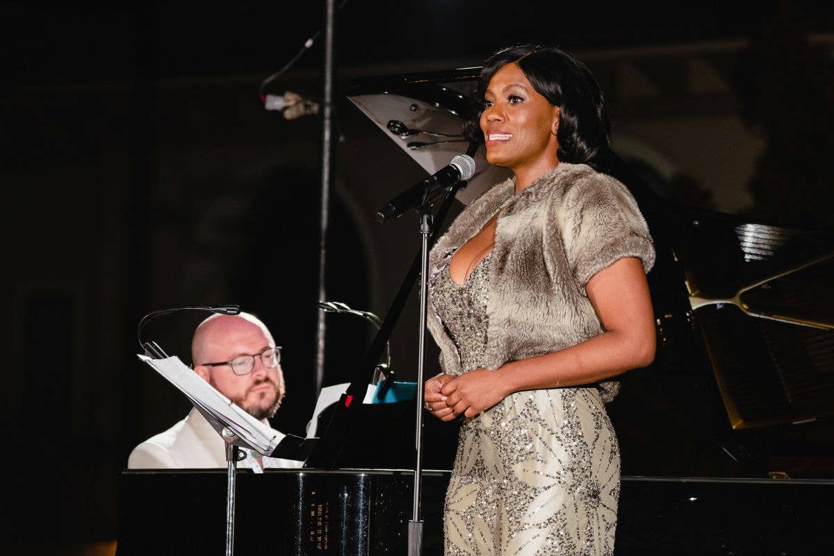 Woman in a sparkly dress sings at a microphone beside a grand piano, with a seated pianist in the background.