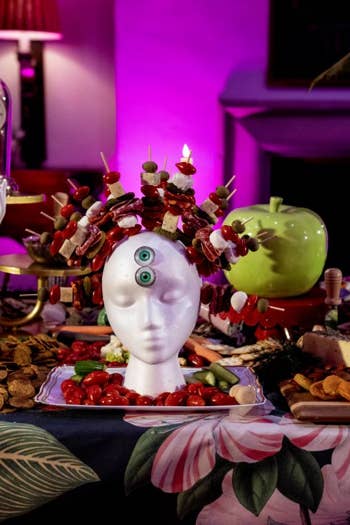 Surreal festive table scene with decorated head sculpture, candles, fruits, and purple-lit clock in the background