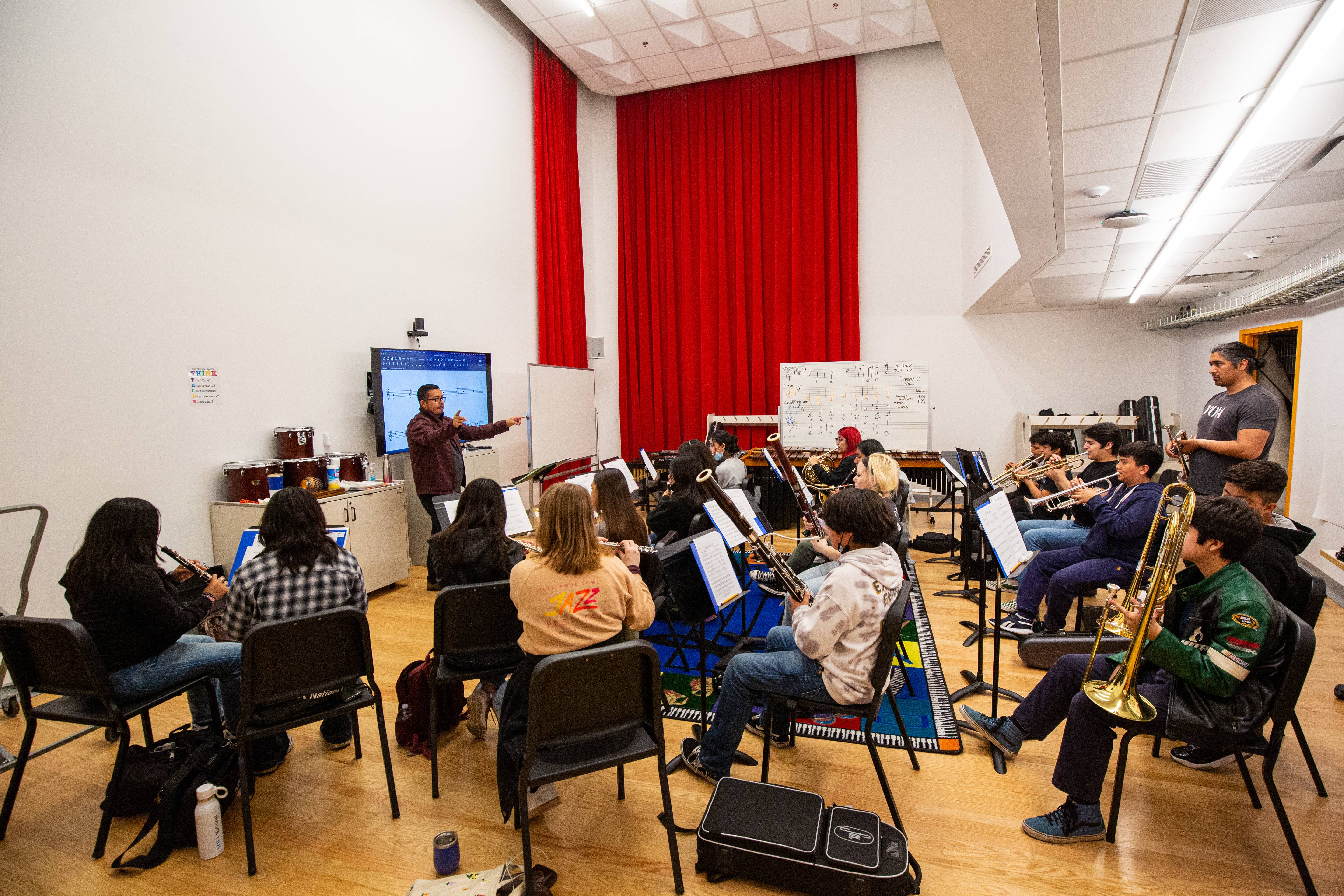 Music classroom with a teacher conducting a brass and woodwind rehearsal in front of red curtains.