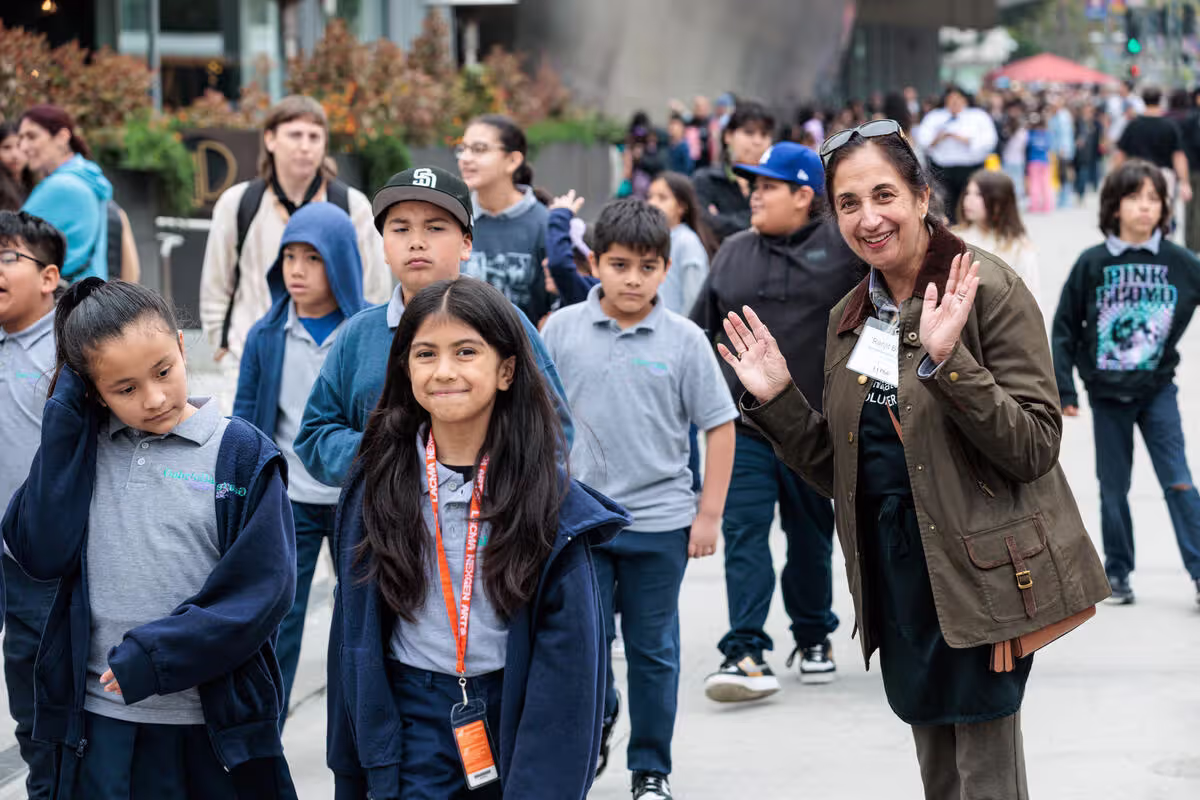 Woman in green jacket waving to students during outdoor event on a busy walkway