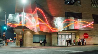 Neon light sculpture wraps a the Roy and Edna Disney CalArts Theater building exterior with people standing outside at night.