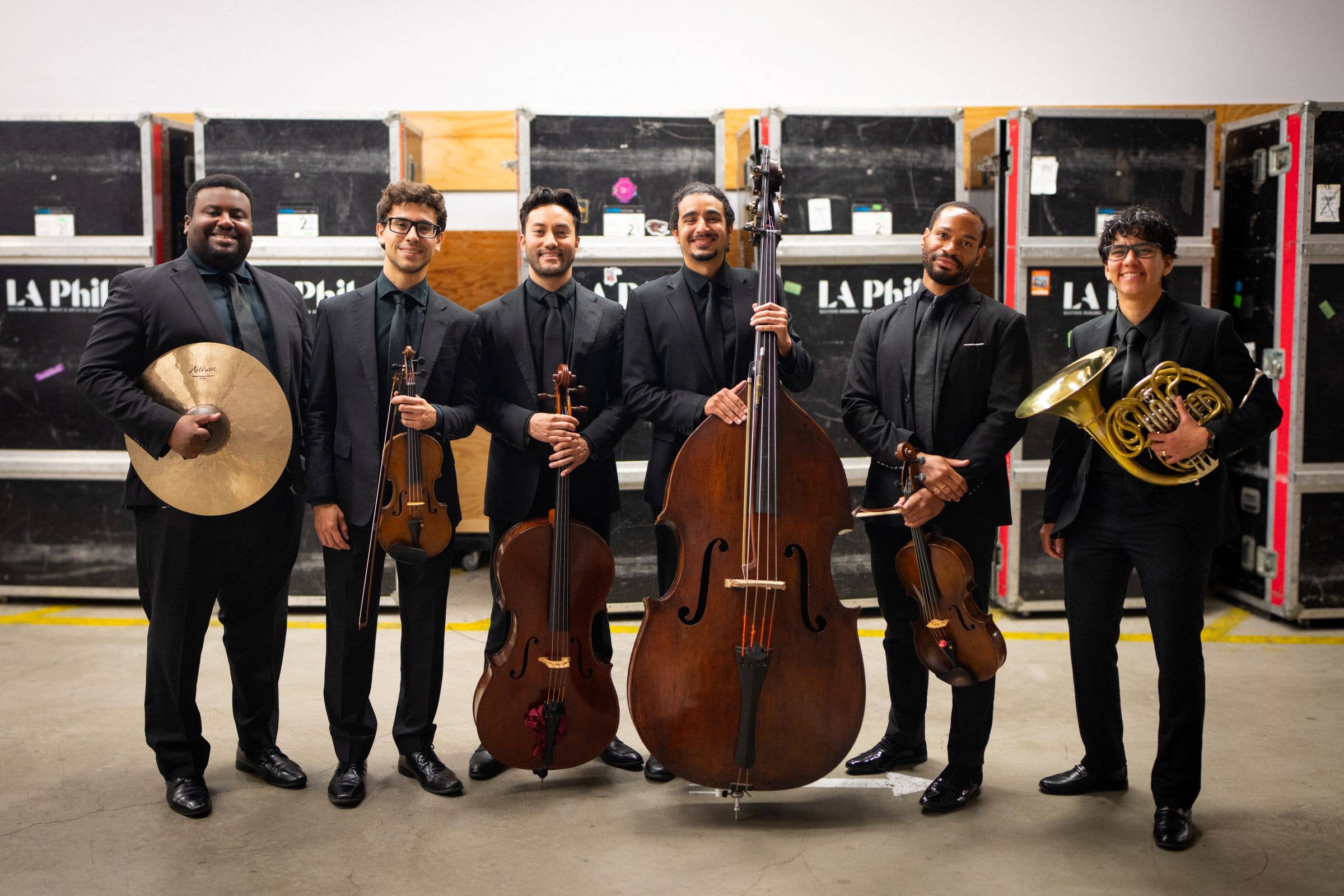 Six musicians in black suits pose with instruments, including cello, violin, gong, trumpet, and a drum in a studio.