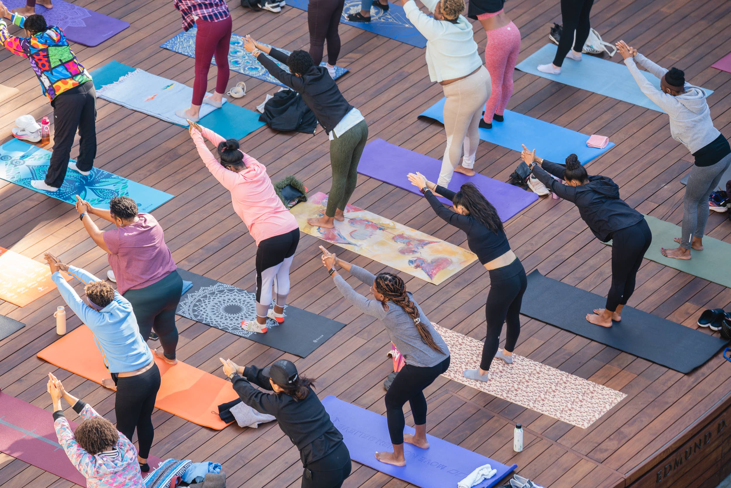 Group yoga class on blue and purple mats, participants stretching with hands overhead on a wooden deck