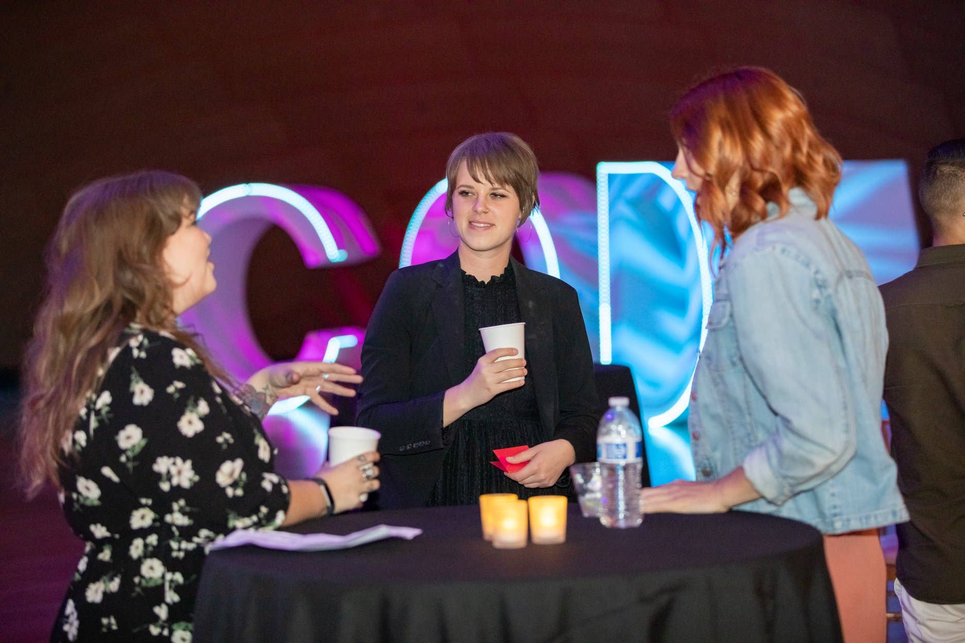 Conference panel with three people chatting at a table, woman holding a cup, colorful neon backdrop.