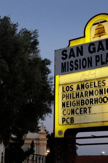 Yellow marquee sign reading “San Gabriel Mission Playhouse” with event details, framed by trees and dusk sky