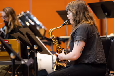 Musician seated with gold saxophone in a rehearsal room, facing sheet music and microphones on stands.