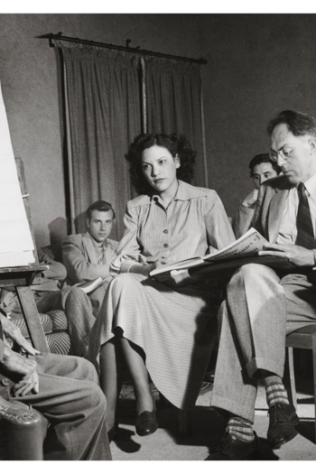 Men and women seated in a meeting, one man at an easel with sheet music background, others reading papers.