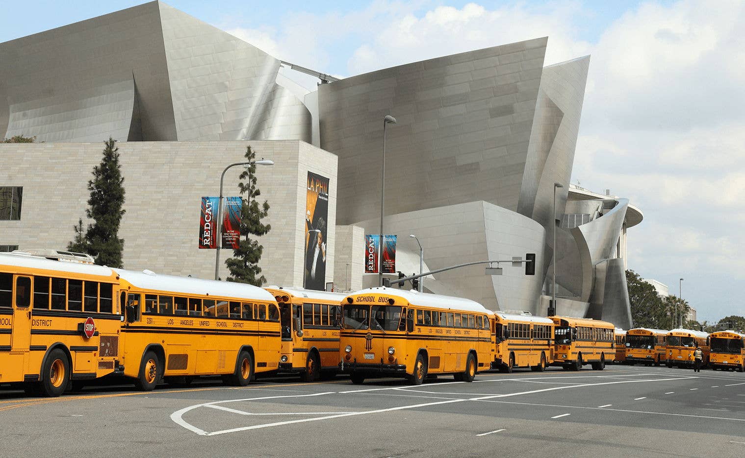 Yellow school buses parked in front of a modern gray civic building with banners at curbside.