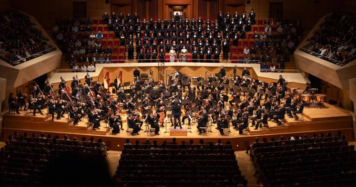 Orchestra and choir performing together on a concert hall stage with musicians seated and standing in rows.