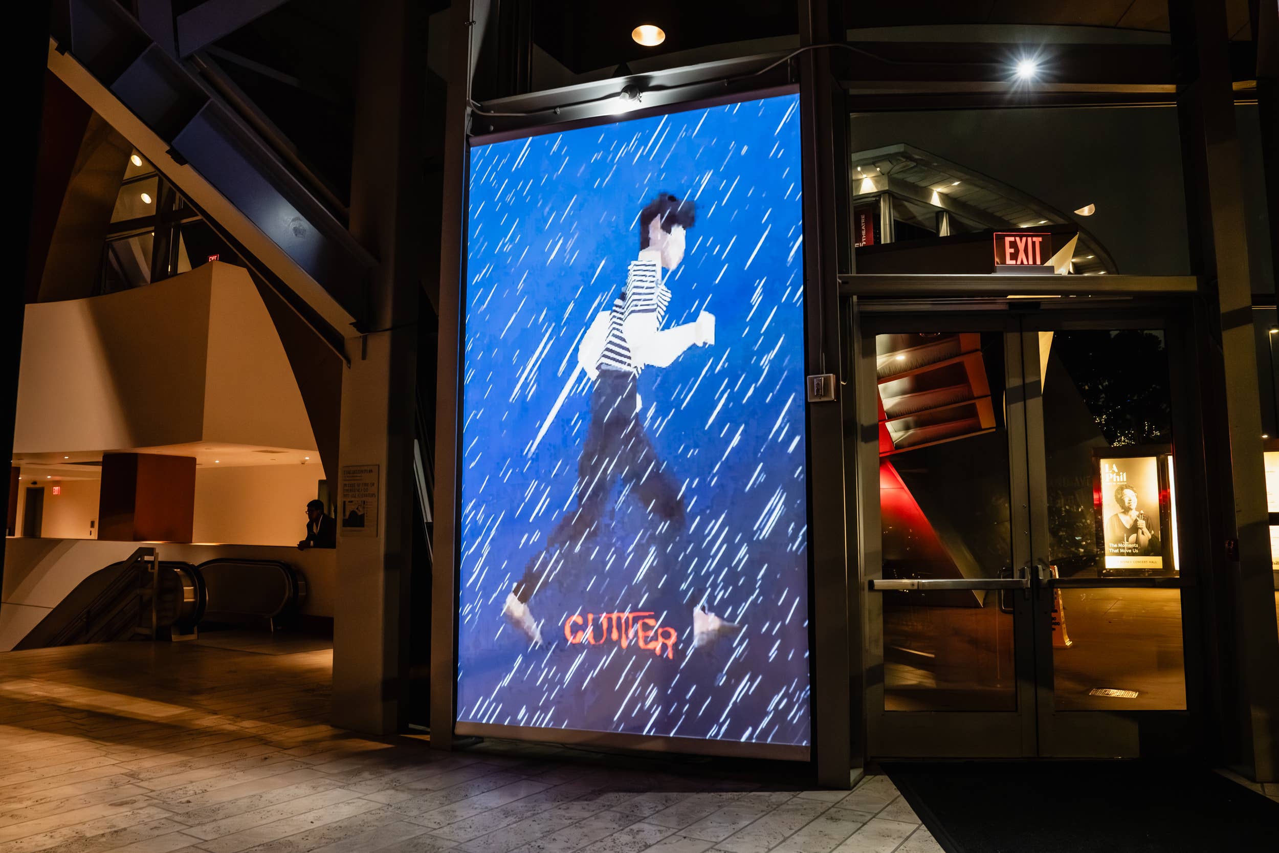 Indoor venue entrance with digital screen showing rain and a character holding an umbrella; exit sign above doors.