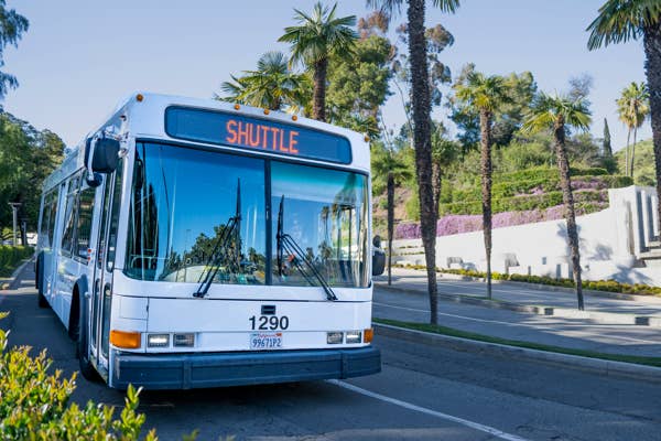 Un autobús lanzadera con el letrero «Shuttle» se detuvo en una carretera soleada, rodeada de palmeras y arbustos bien cuidados.
