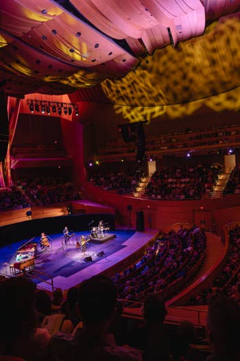 Concert hall stage with colorful lighting, musicians performing, and packed audience seated in curved balconies
