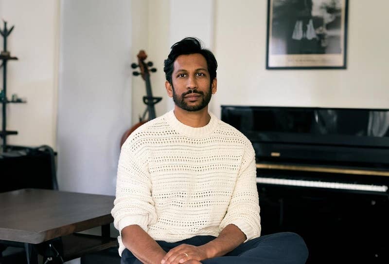 Man with dark hair and beard wearing a white sweater, sitting in a room beside a piano and table.