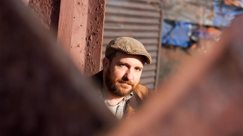 Bearded man wearing a tweed flat cap and checkered shirt, looking at the camera through window blur.