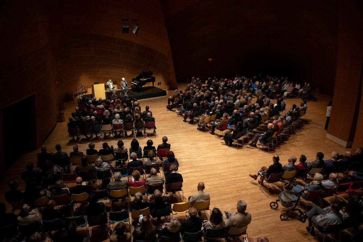 Audience seated in a concert hall as musicians perform on stage with piano and speakers overhead.
