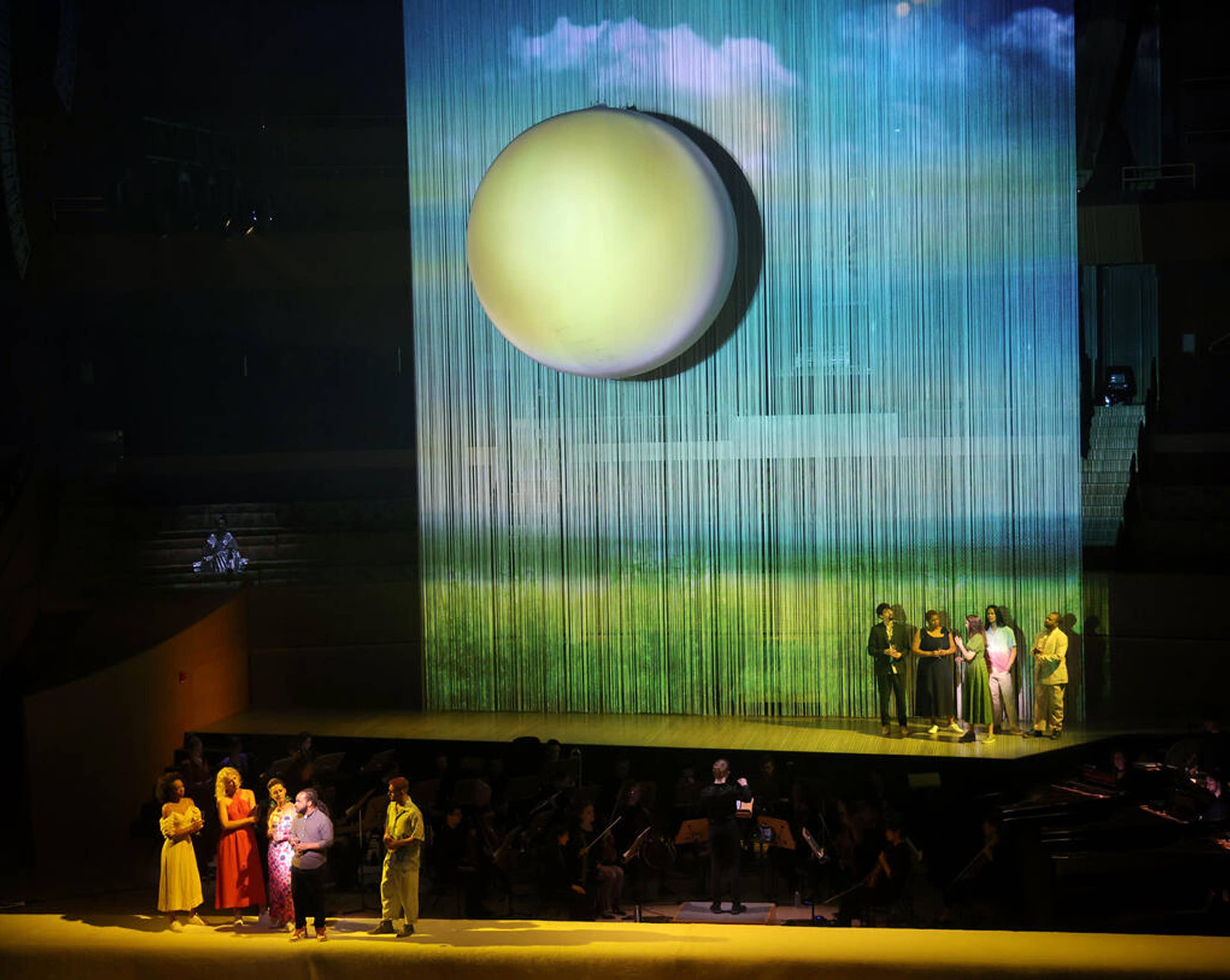 Stage performers stand before a large glowing sphere on a blue, vertical light curtain screen.