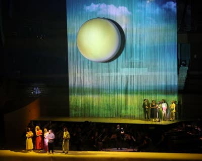 Stage performers stand before a large glowing sphere on a blue, vertical light curtain screen.