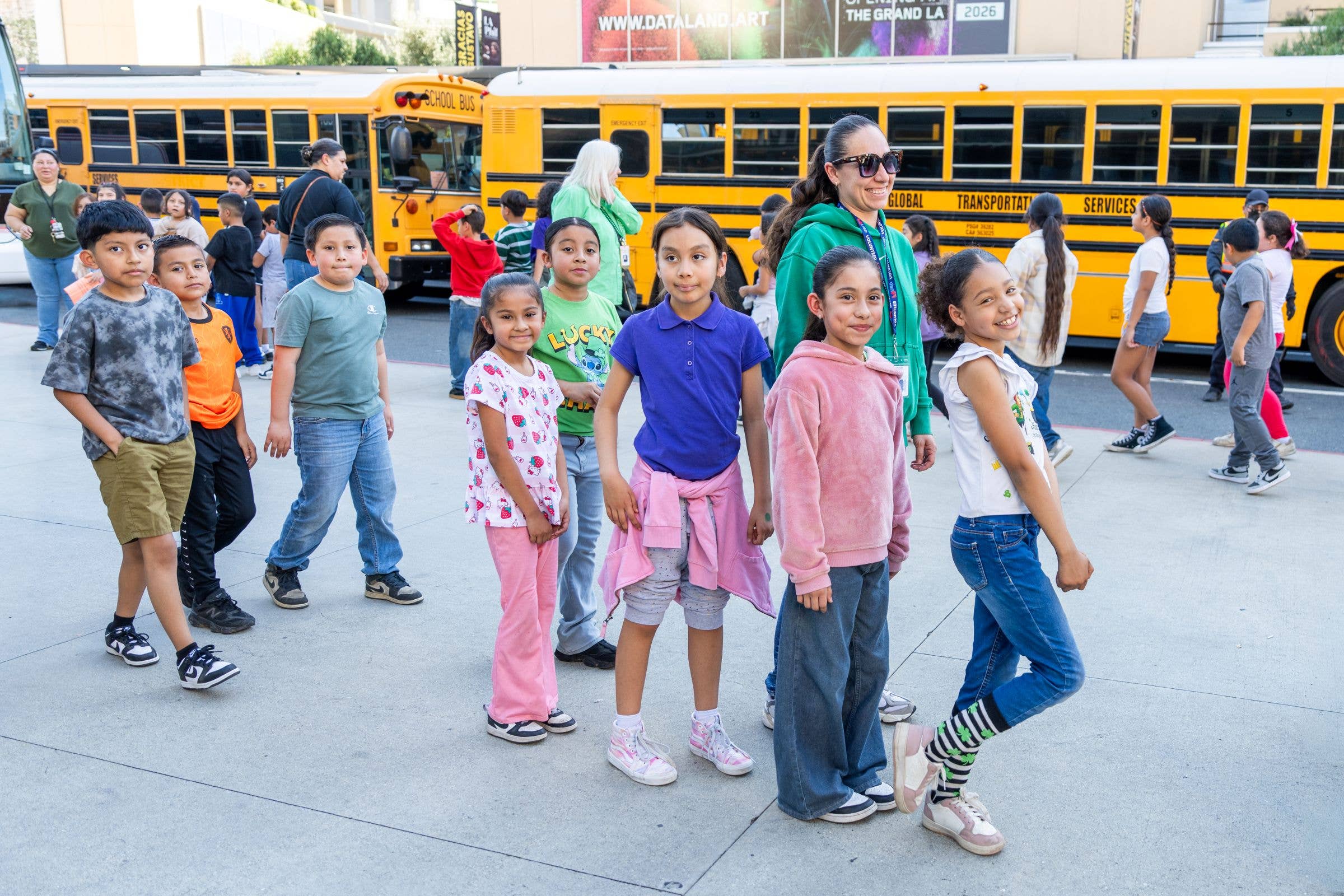 Smiling children and staff walk in front of a yellow school bus outside a building during school pickup.