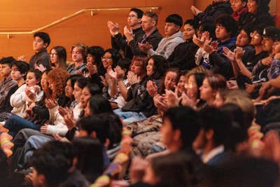 Students in an auditorium clap and cheer with raised hands during a school event.