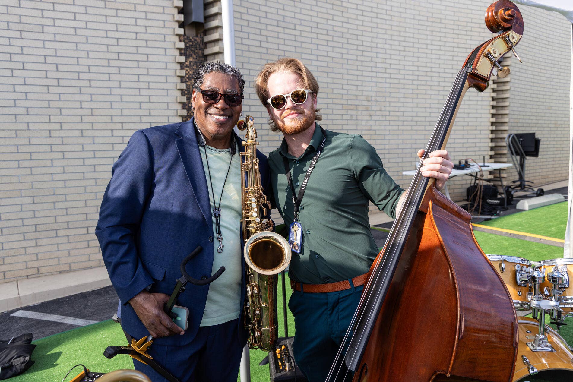Two musicians in sunglasses pose with a saxophone and a large upright bass outside a brick building.