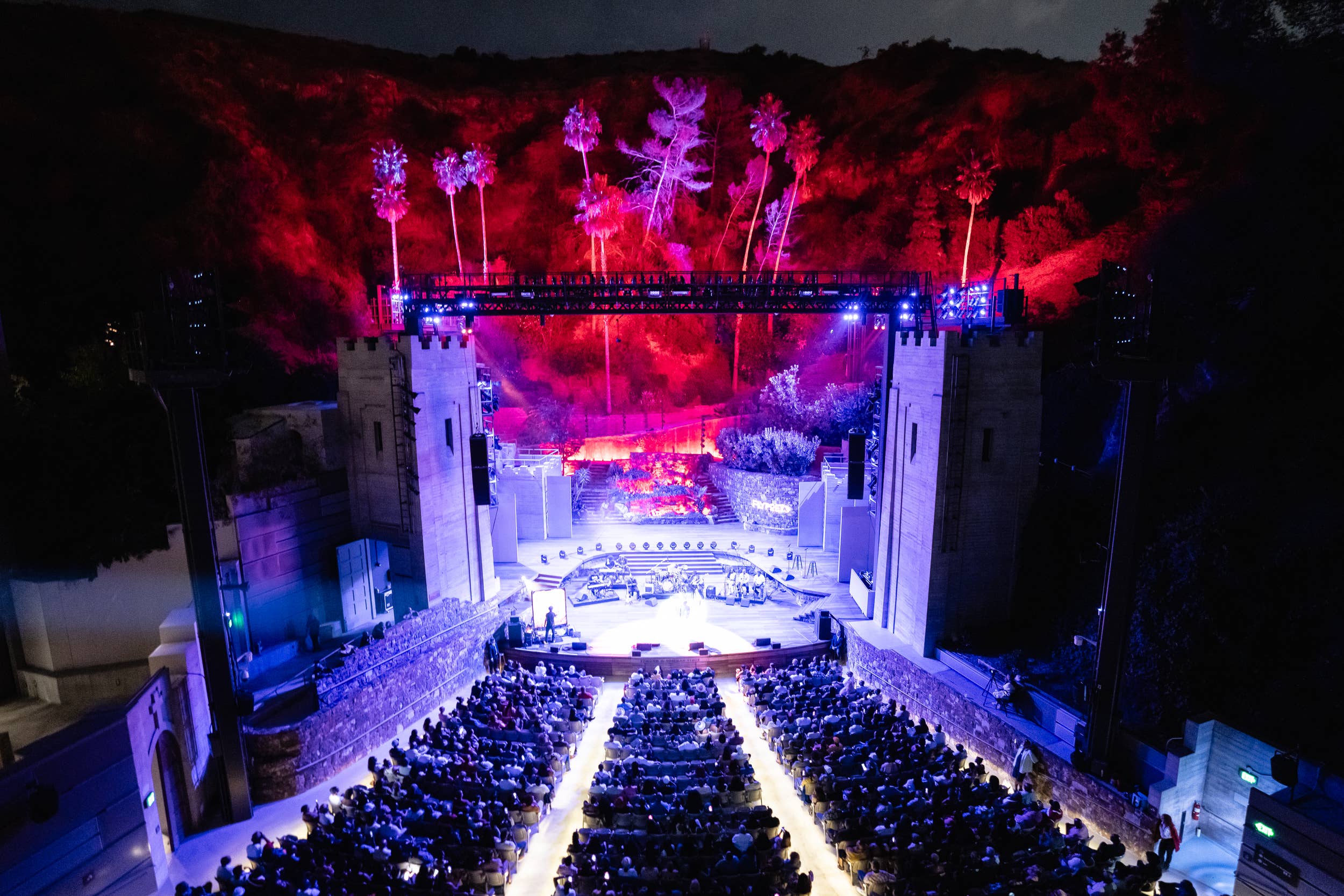 Outdoor evening concert stage with audience, purple and red lighting, and glowing fireworks-style pyrotechnics over hills