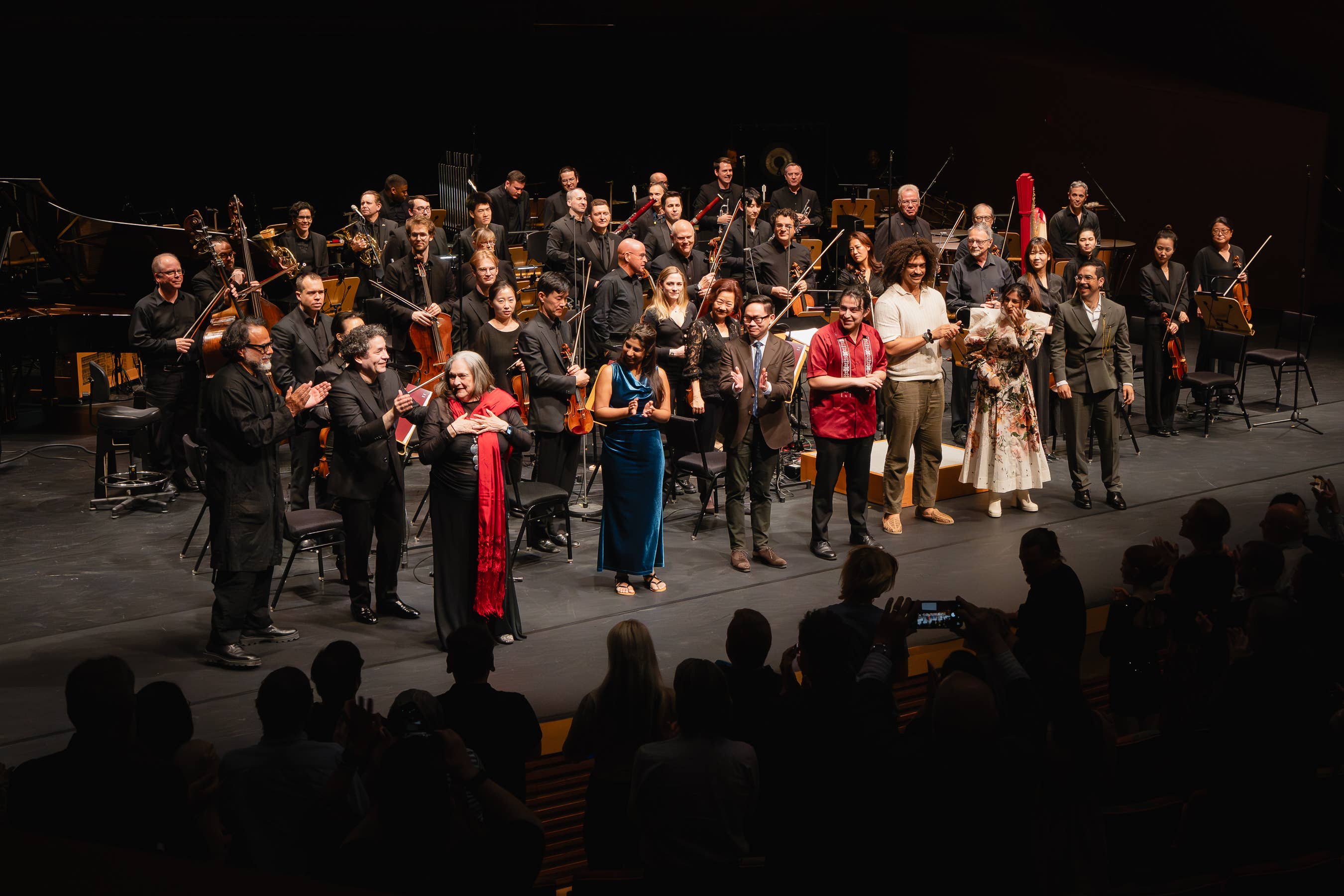 Orchestra and soloists in formal attire standing on stage with instruments, smiling during a concert after-performance photo