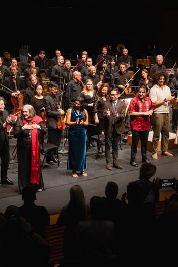 Orchestra and soloists in formal attire standing on stage with instruments, smiling during a concert after-performance photo