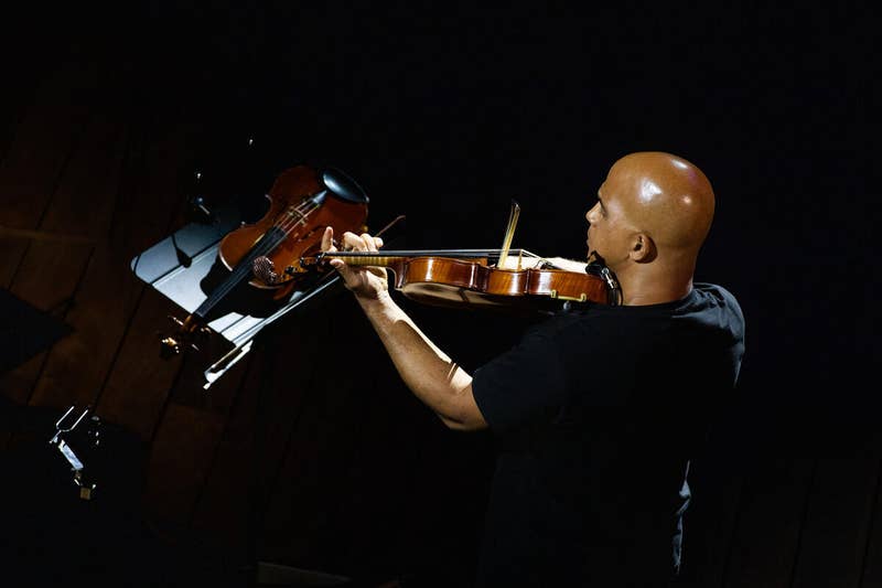Two musicians play instruments on stage under dark lighting: violin and guitar with spotlight.