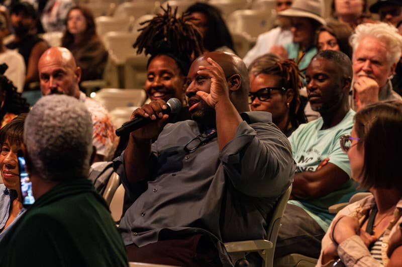 Man in suit speaks into a handheld microphone, gesturing, as audience members sit in rows.