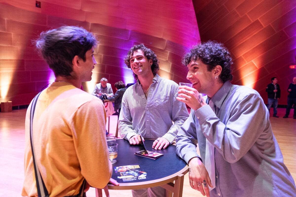 Three men chat and laugh at a round table during an event, with colorful pink and purple lighting in a hall.