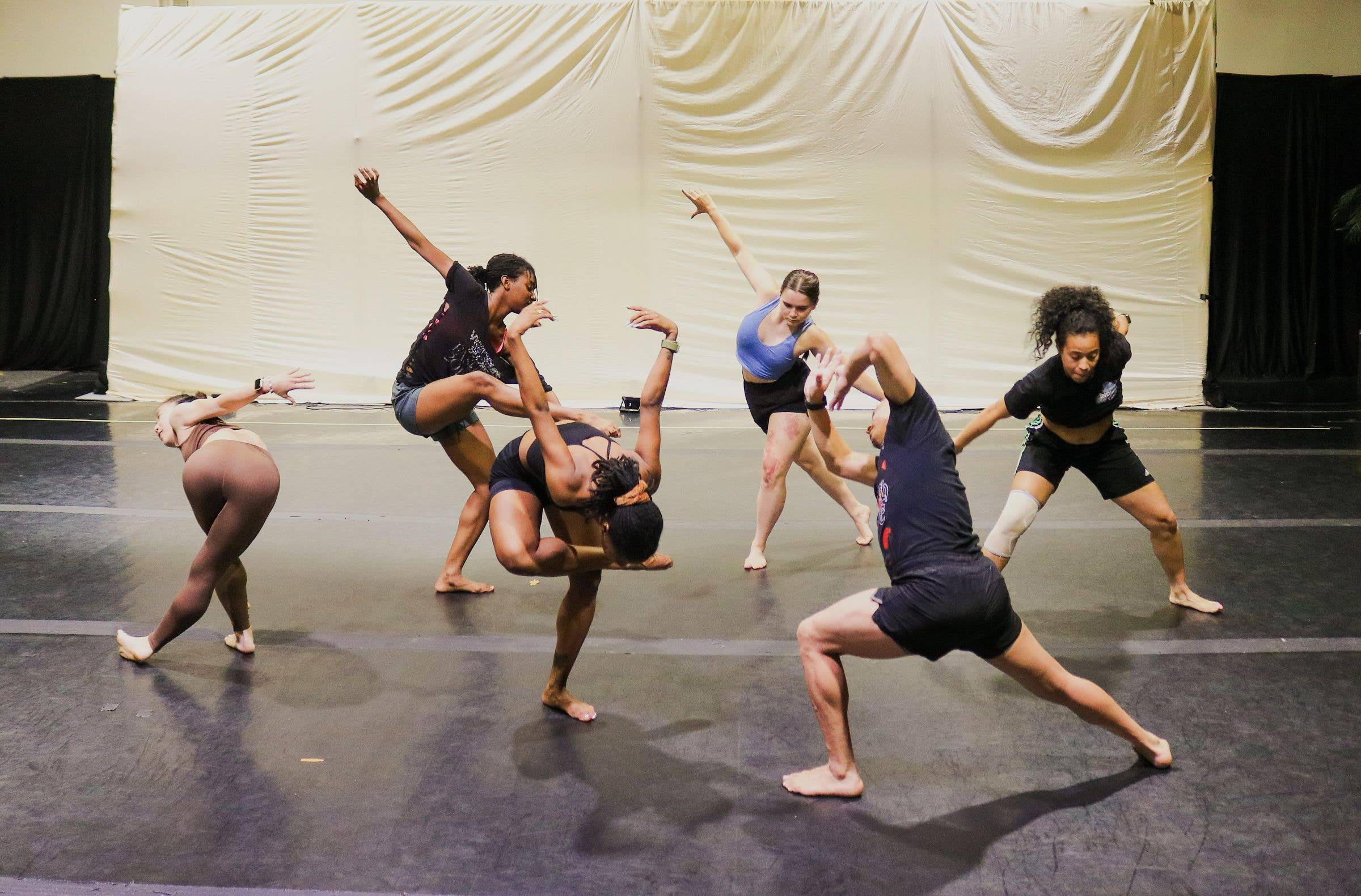 Dancers perform acrobatics on a gym floor, lifting and balancing with arms raised in front of a white backdrop.