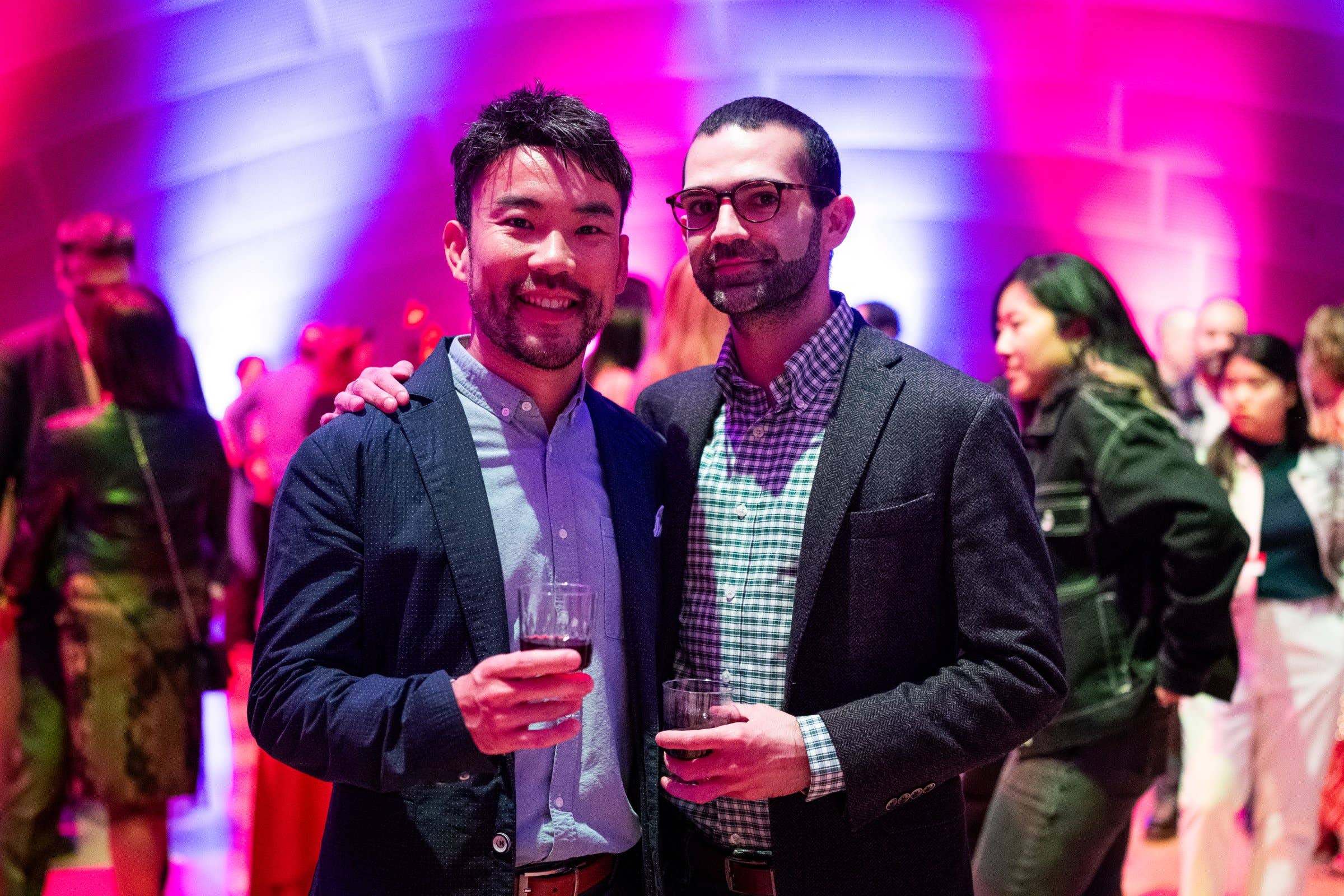 Two men smiling and holding drinks at a party under colorful pink and purple stage lights