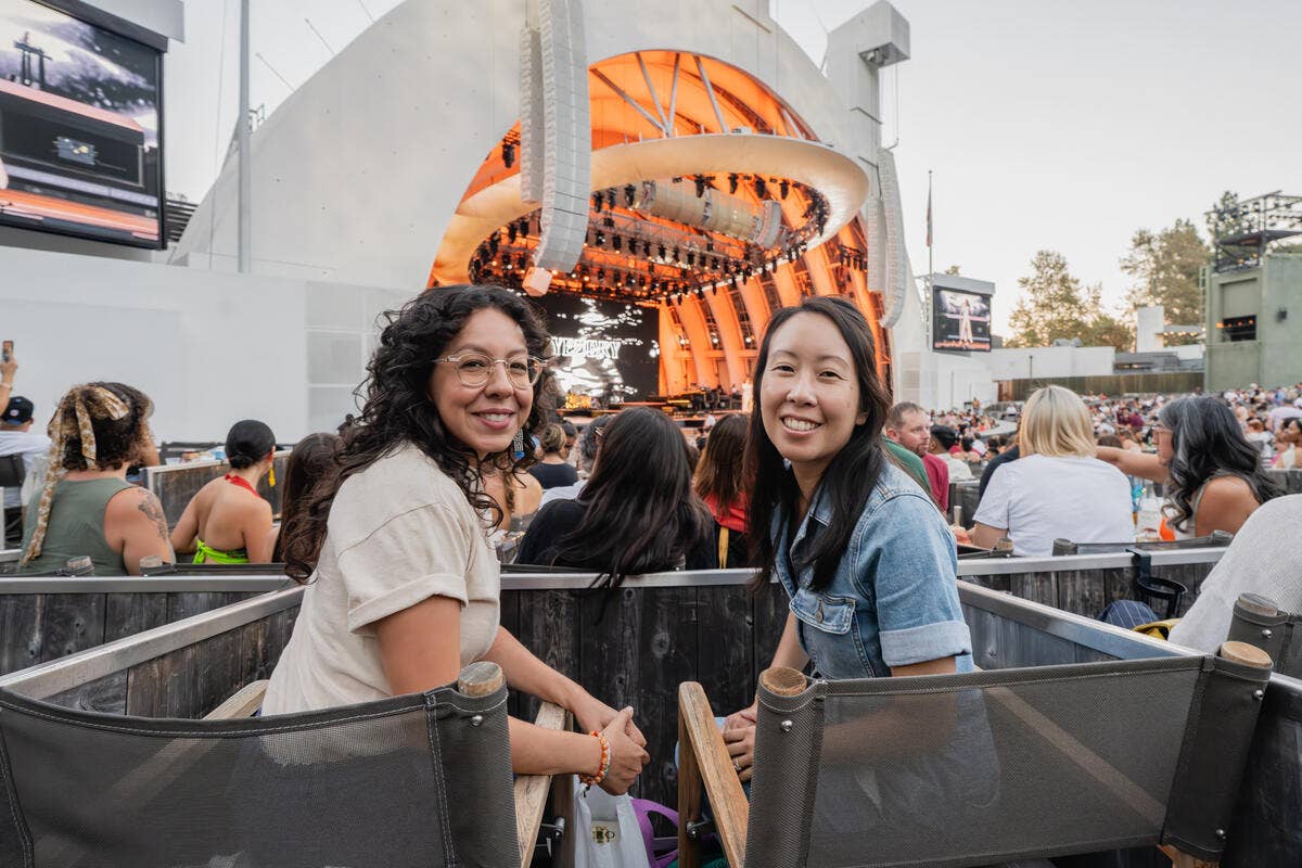 Two women smiling at an outdoor concert, with an orange-lit stage in the background and crowds behind them.