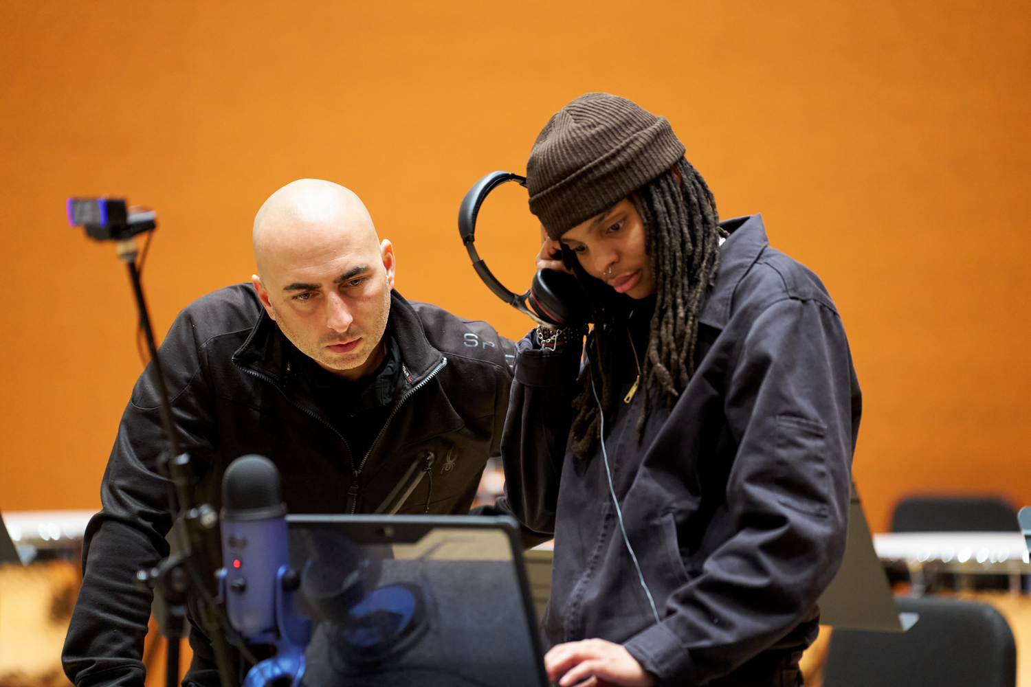 Two men, one bald with focused expression and another wearing a beanie and headphones at a laptop.