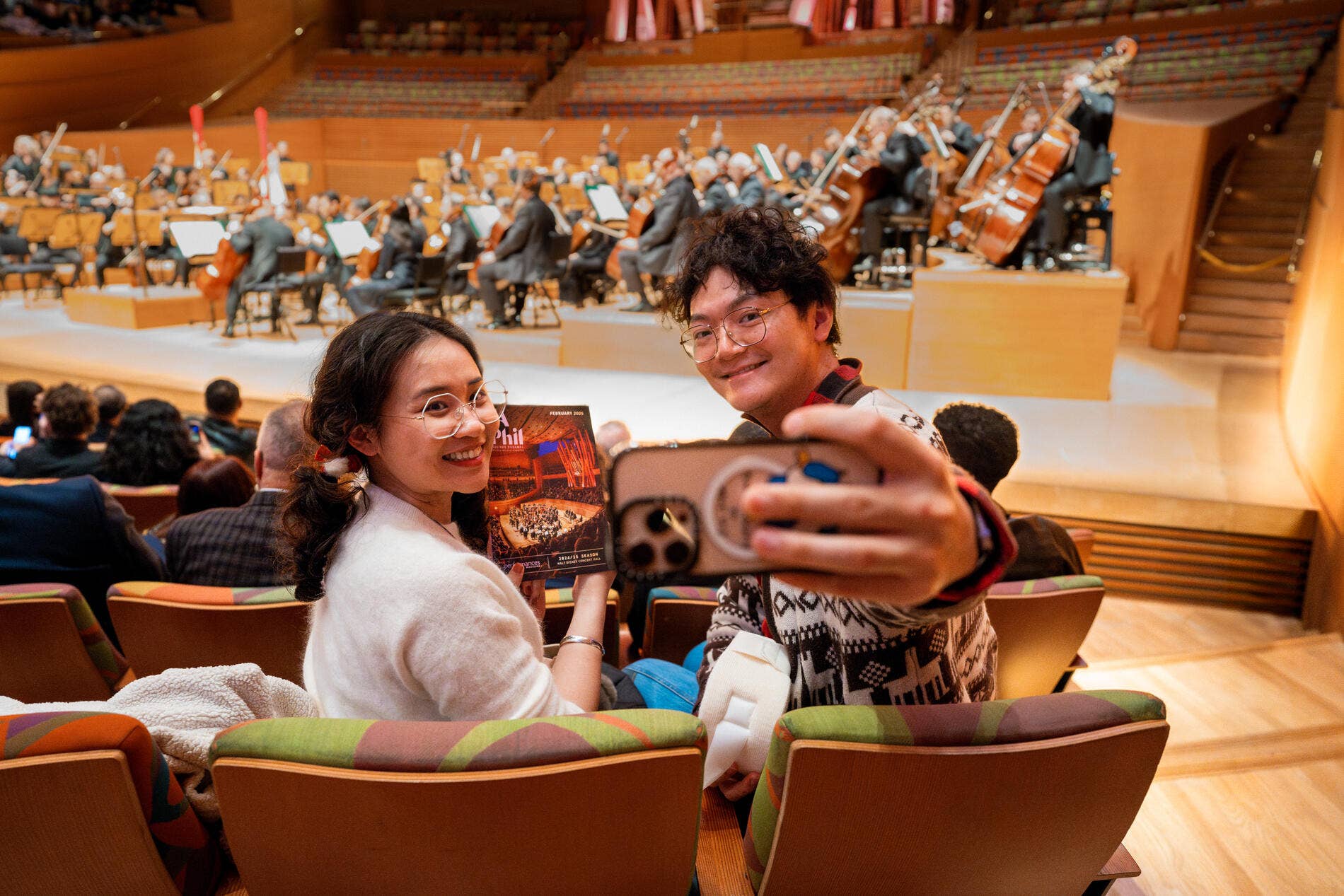 Smiling couple takes a selfie in a theater while an orchestra performs on stage.