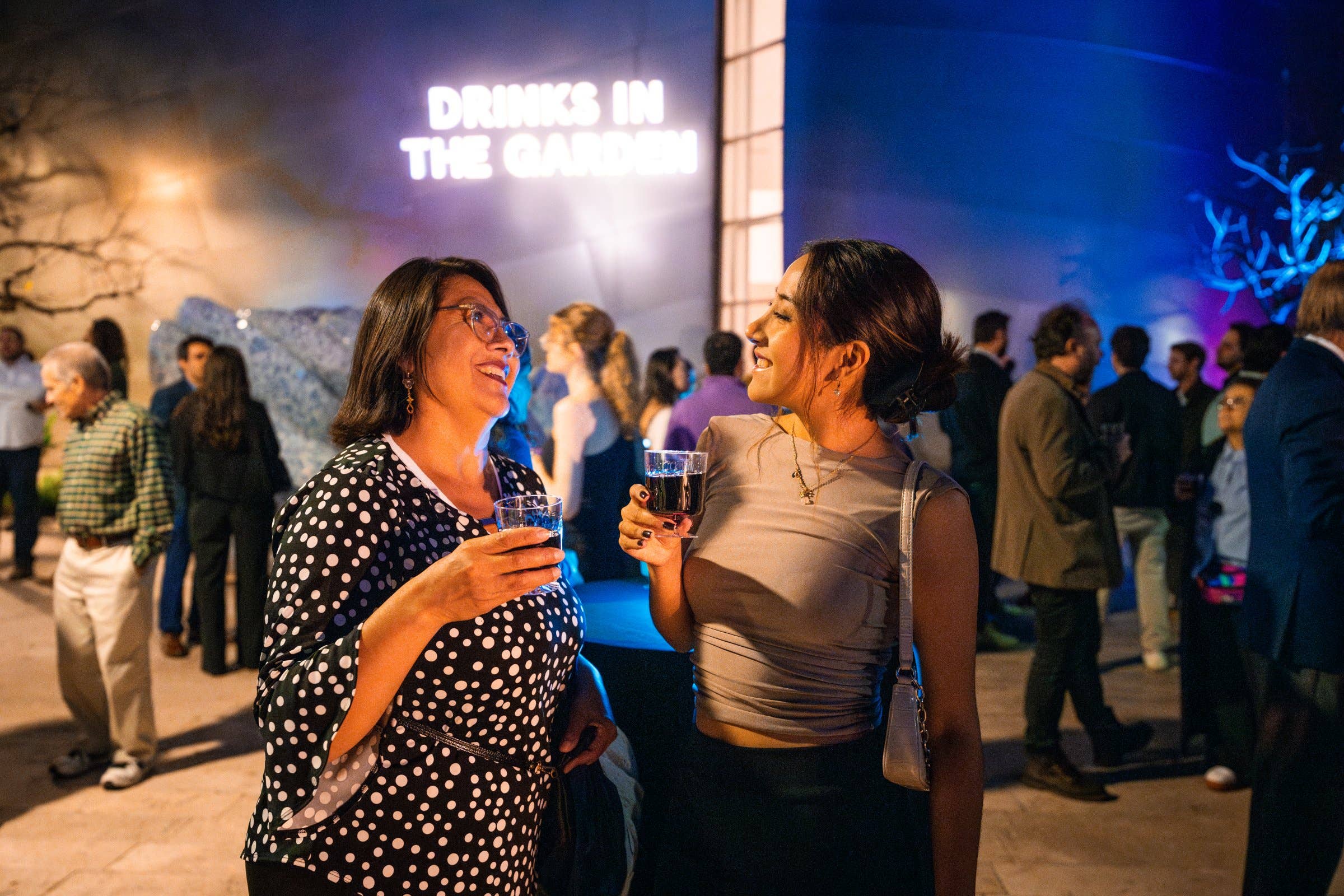Two women smile and clink drinks at a nighttime party with a “Drinks in the garden” neon sign behind them