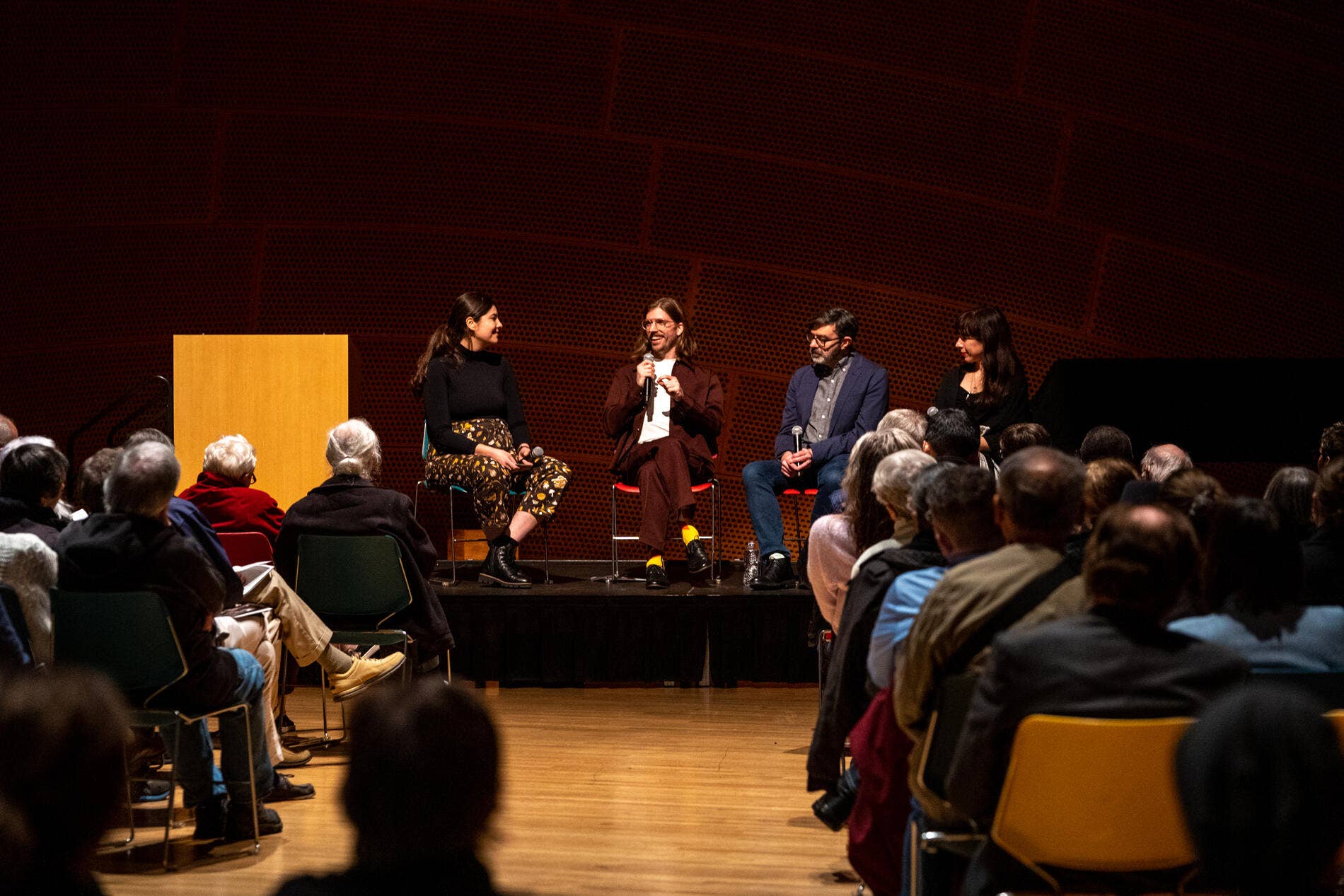 Panel discussion on stage with four speakers seated, one holding a microphone, audience in foreground.