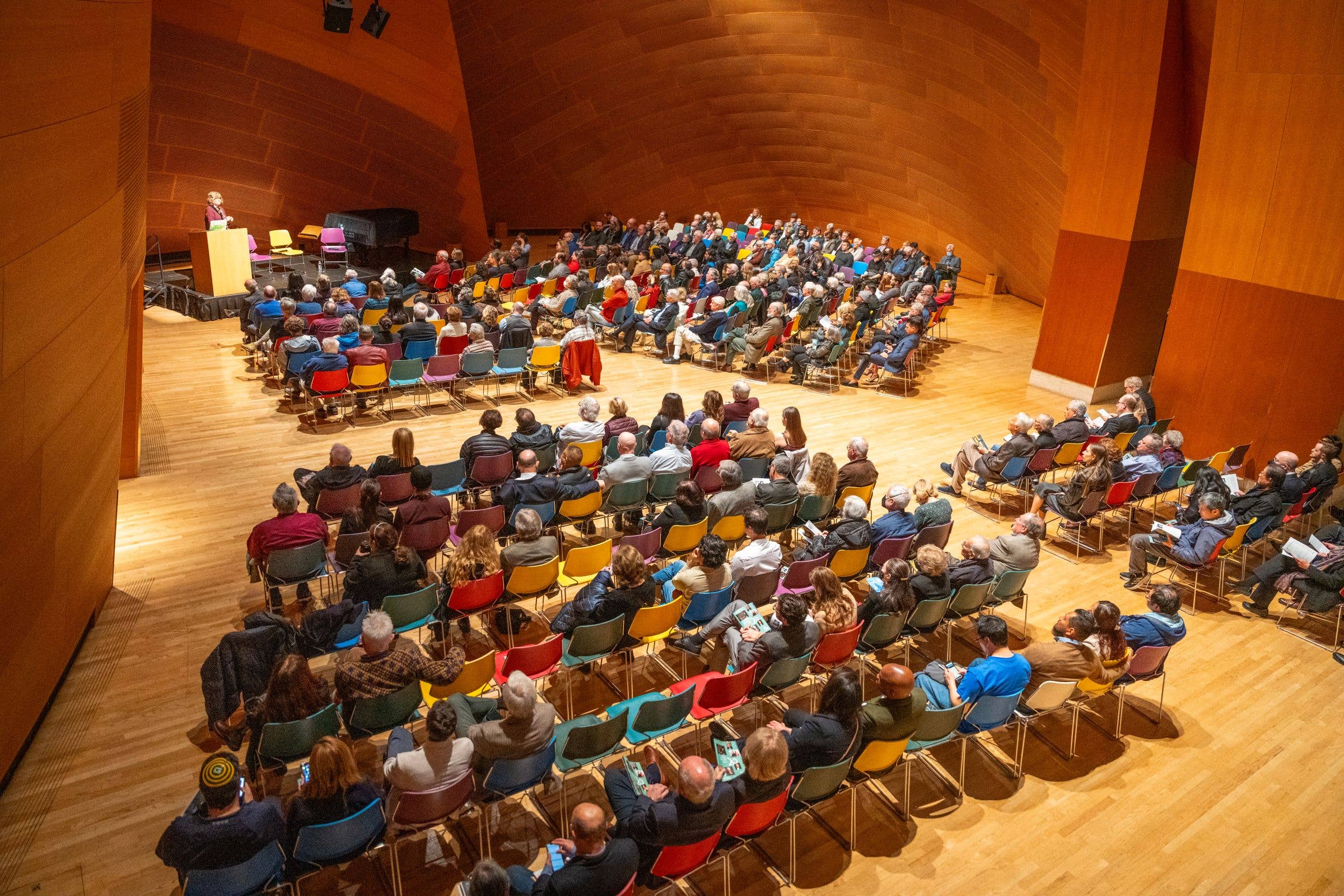 Audience seated in a large concert hall as a speaker presents from a podium on stage