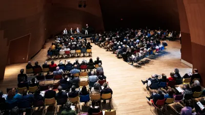Audience seated in a concert hall as speakers stand on stage with microphones and music stands