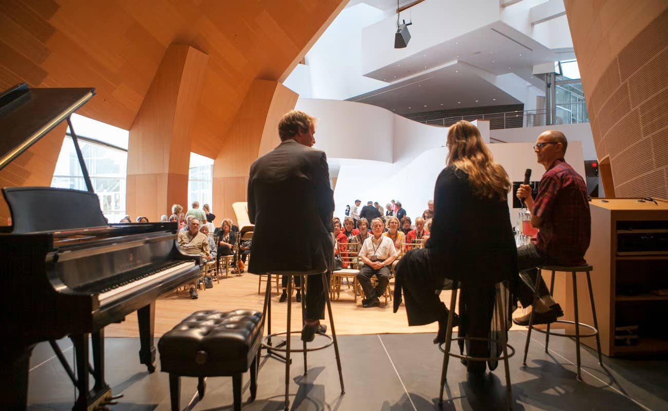 Speakers perform at a concert hall with an audience seated; grand piano and musicians stand in foreground.