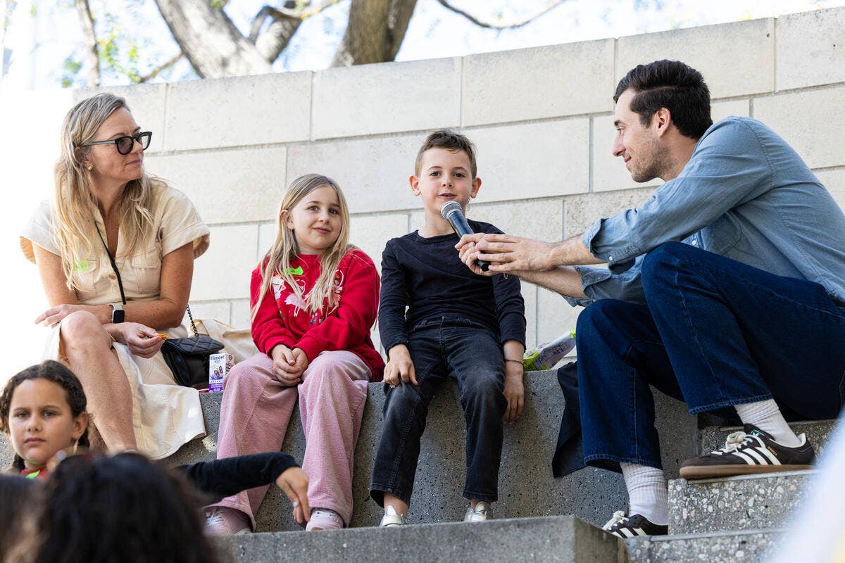 Man kneels outdoors holding a microphone, talking to three children sitting on concrete steps near a woman