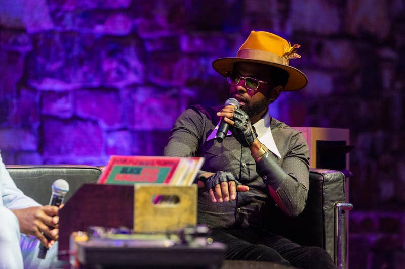 Un hombre con un sombrero fedora de color beige, traje y gafas de sol habla por un micrófono en un escenario con iluminación morada.