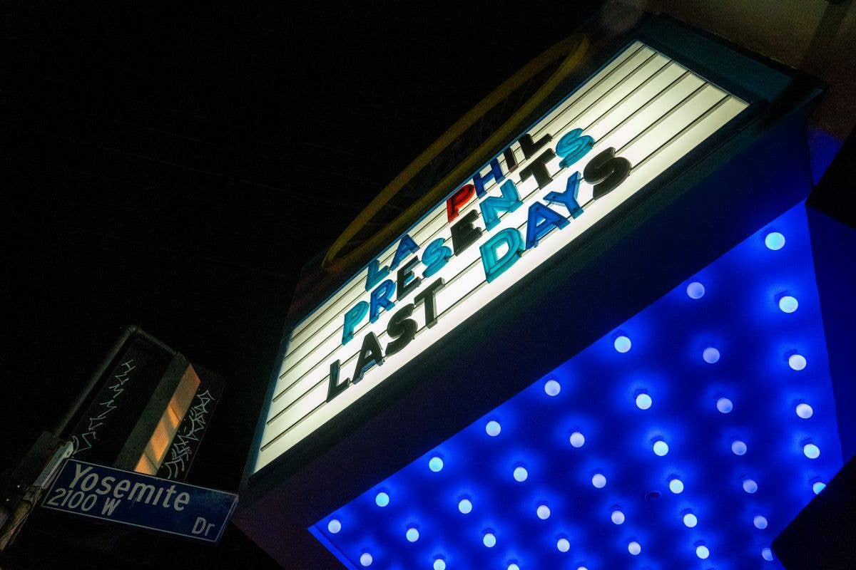 Un letrero de neón que dice «LA PHIL PRESENTS LAST DAYS» en el escaparate de una tienda, con luces azules intermitentes por la noche.