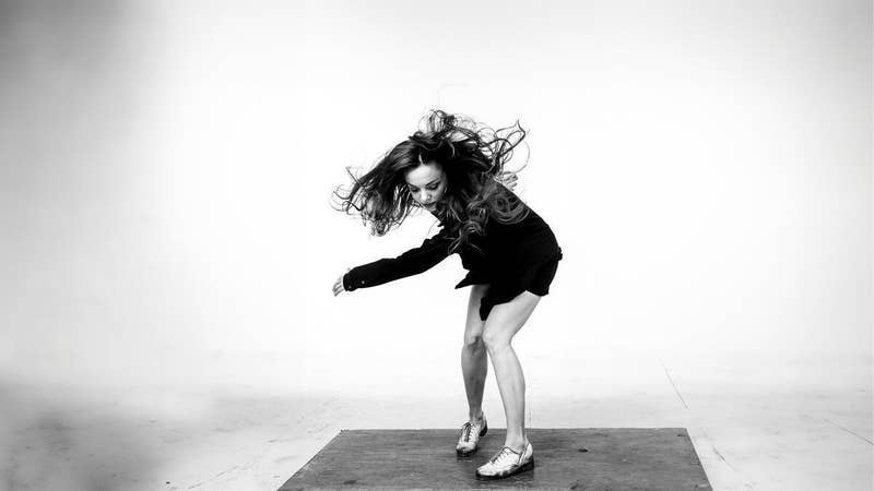 Fotografía en blanco y negro de una mujer bailando en el suelo de un estudio, con el pelo rizado al viento por el movimiento.